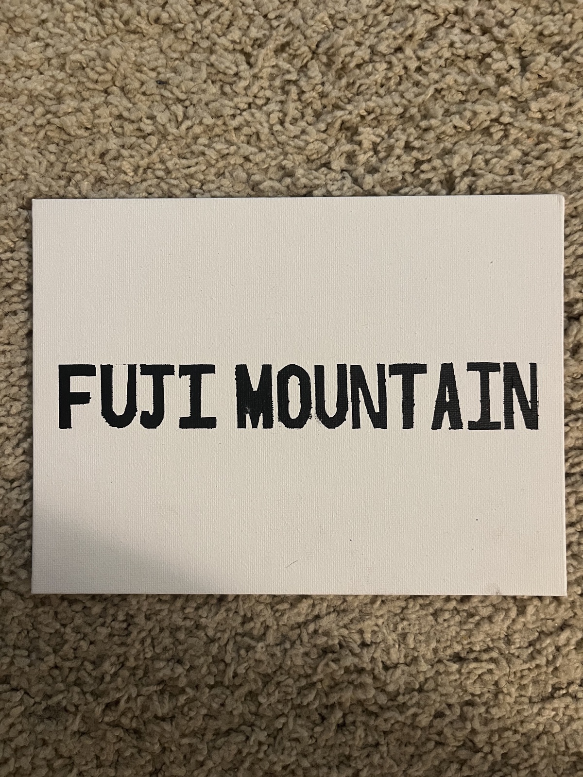 A small white canvas resting on carpet, painted with the words “FUJI MOUNTAIN” in black